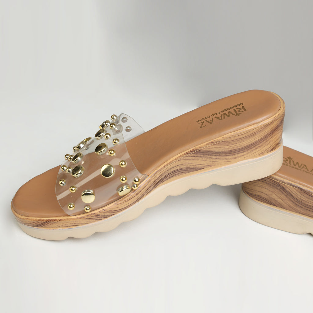 Pair of Riwaaz Nora dark beige gold stud transparent strap wedge slides laid flat with rattan bag and white flowers in background