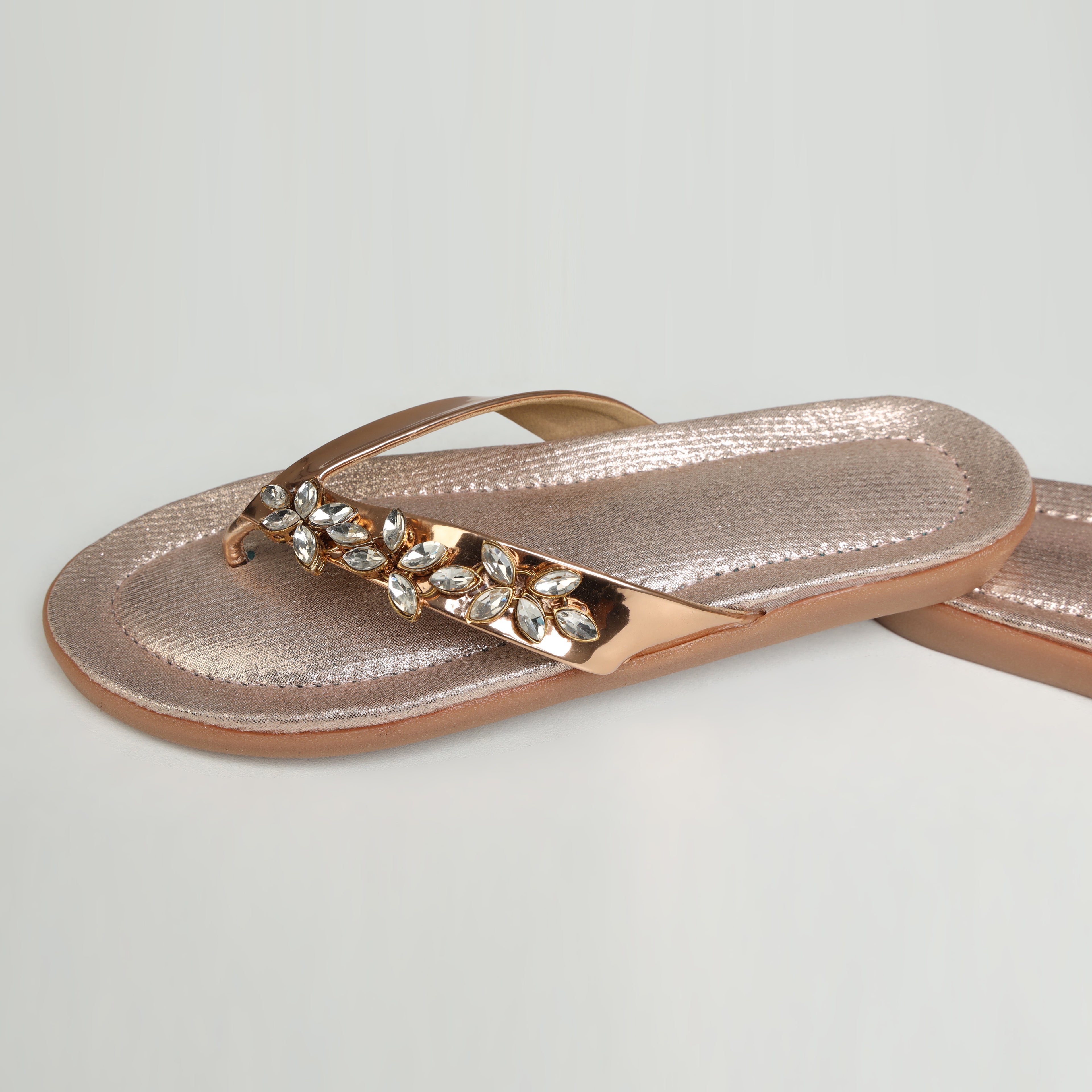 Pair of Riwaaz Ziva rose gold Kundan flip flops styled with white ceramic vase, gold jewellery and white flowers on cream background