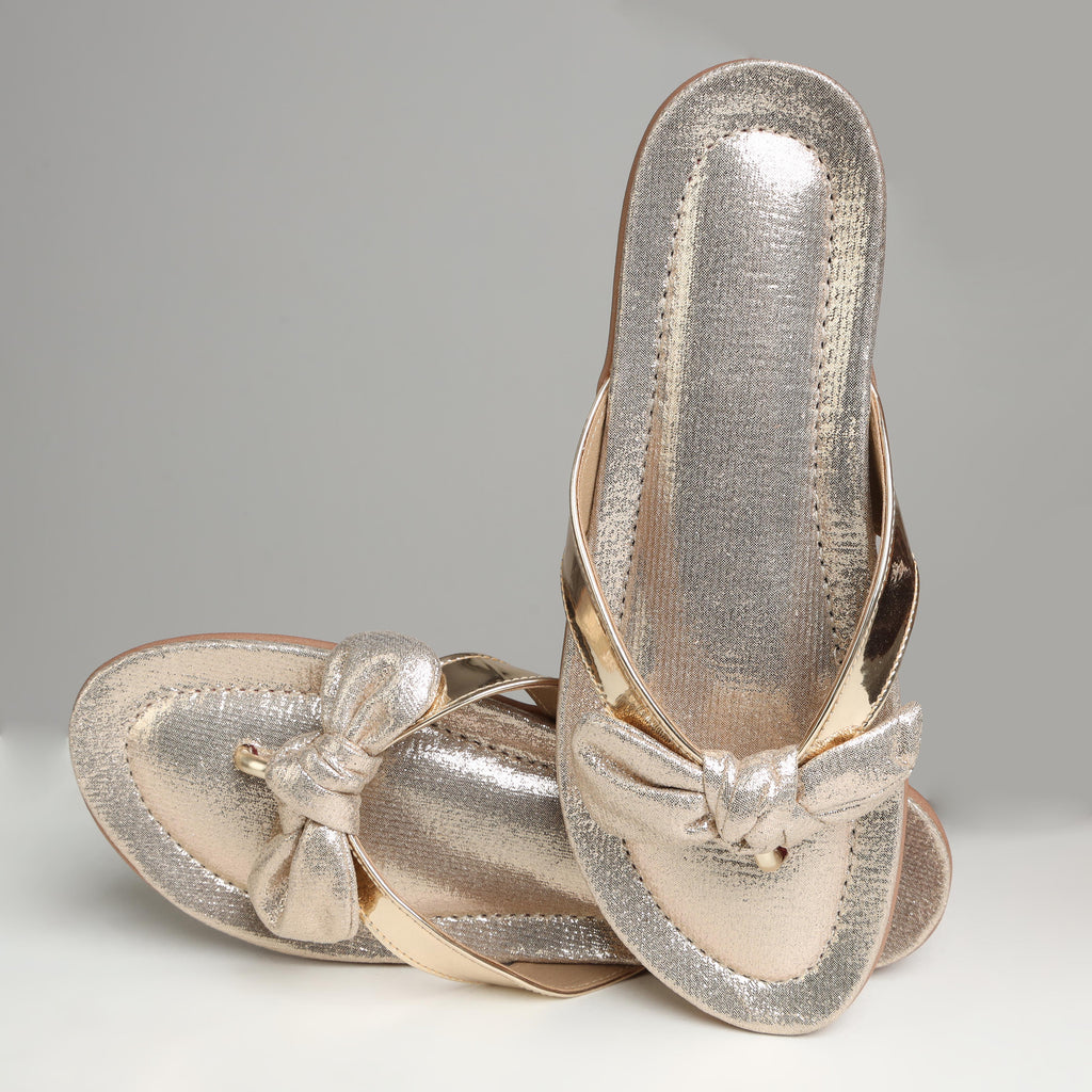 Pair of Riwaaz Misha dull rose gold flip flops on white background – angled top view showing metallic shimmer strap and bow embellishment