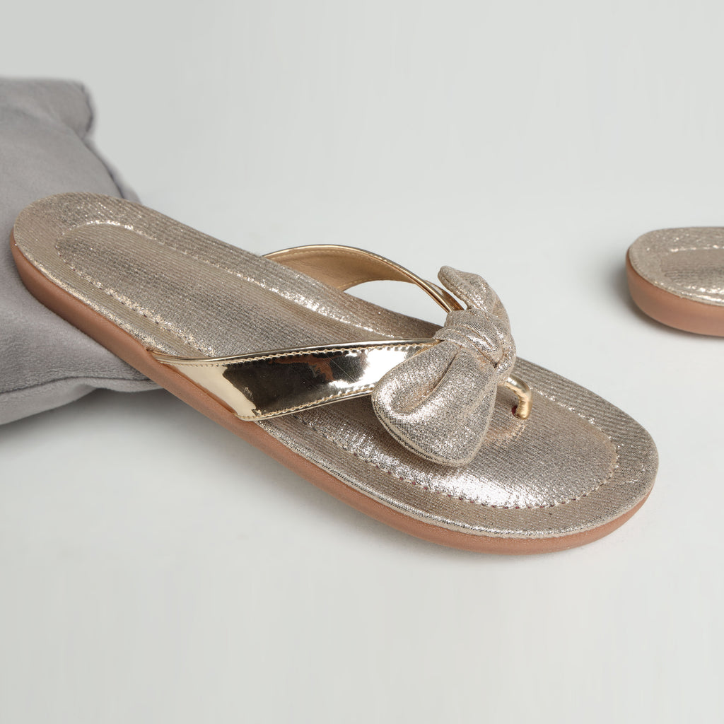 Model wearing Riwaaz Misha dull rose gold mirror work flip flops with white embroidered salwar – close-up foot shot on stone floor with brass pot and flower petals in background