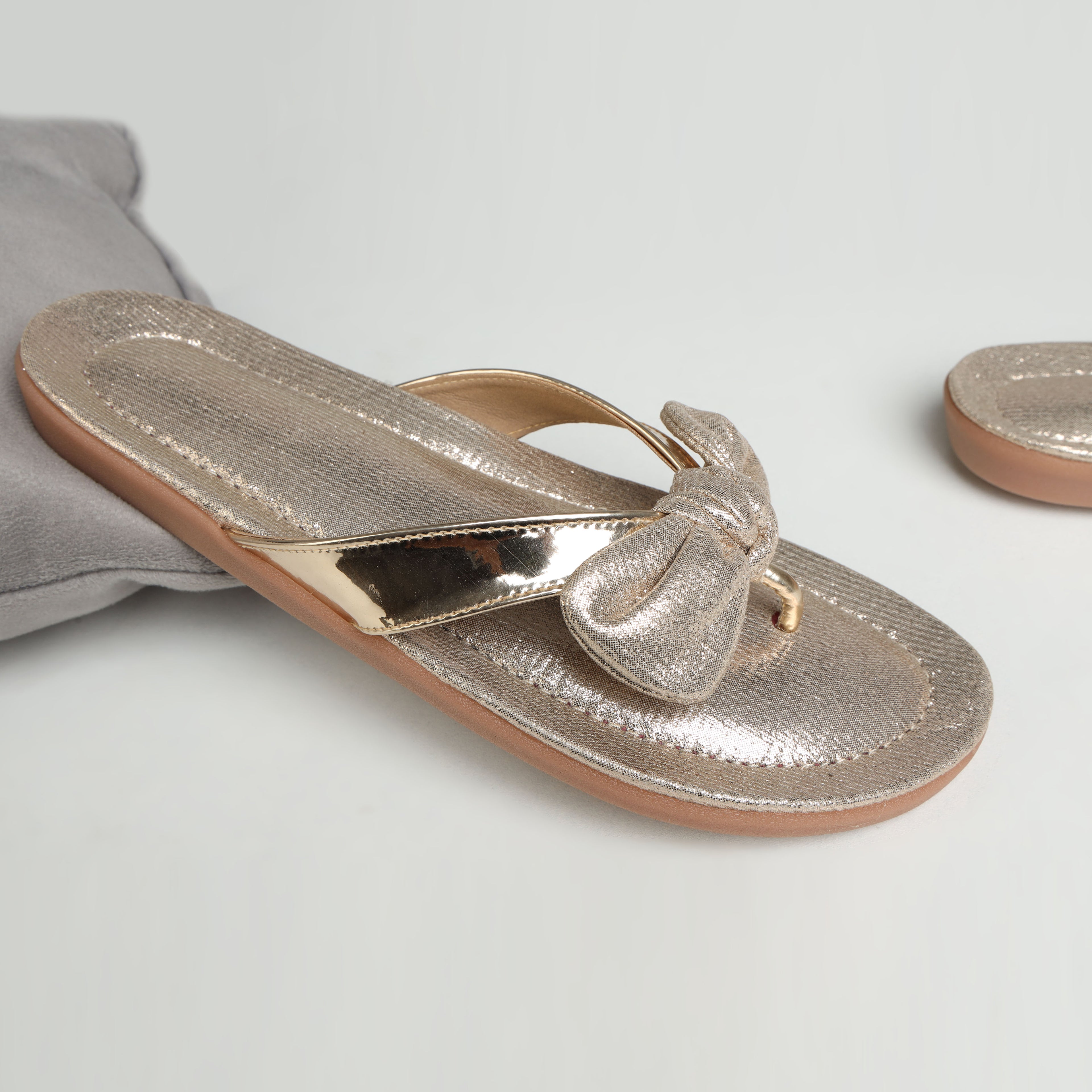 Model wearing Riwaaz Misha dull rose gold mirror work flip flops with white embroidered salwar – close-up foot shot on stone floor with brass pot and flower petals in background