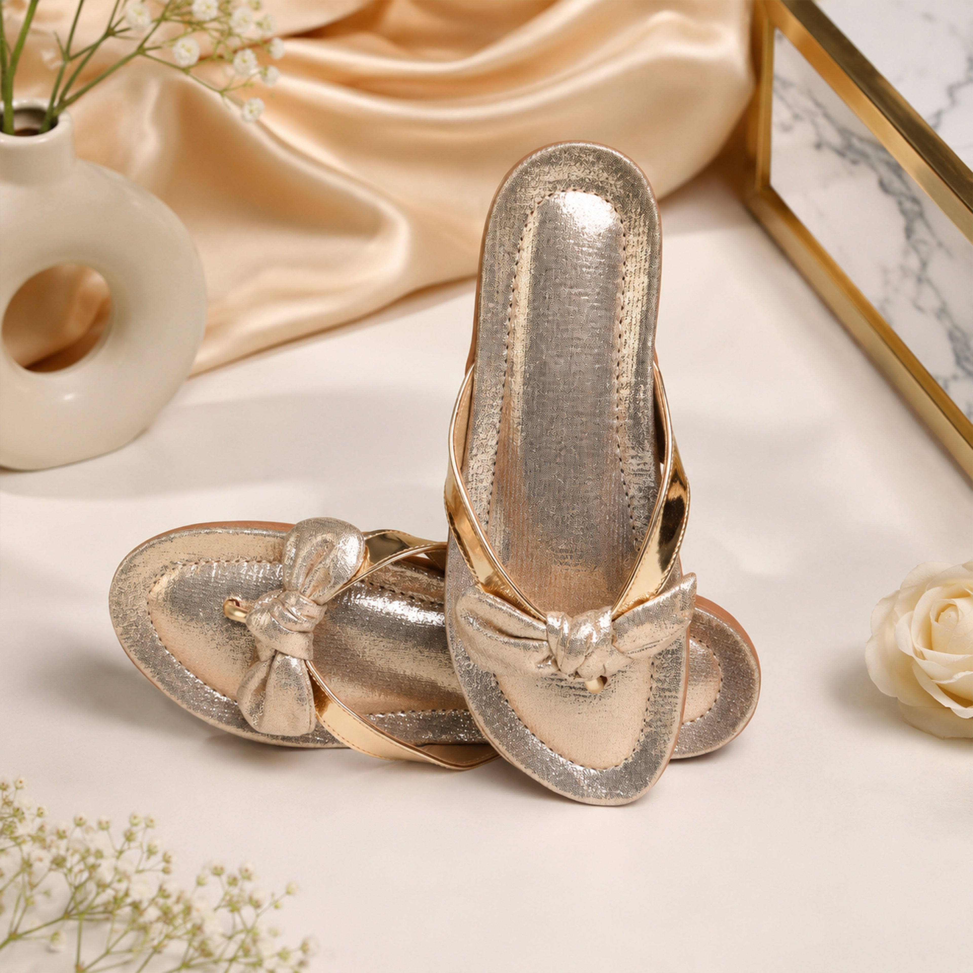 Model seated in cream co-ord set wearing Riwaaz Misha dull rose gold mirror work French knot flip flops – full lifestyle shot with gold drape and white flowers
