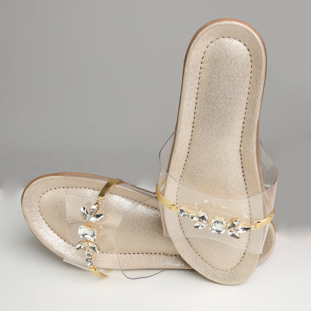 Pair of Riwaaz Ira dull gold crystal transparent strap flats on grey background – angled top view showing full crystal embellishment and gold metal bar detail