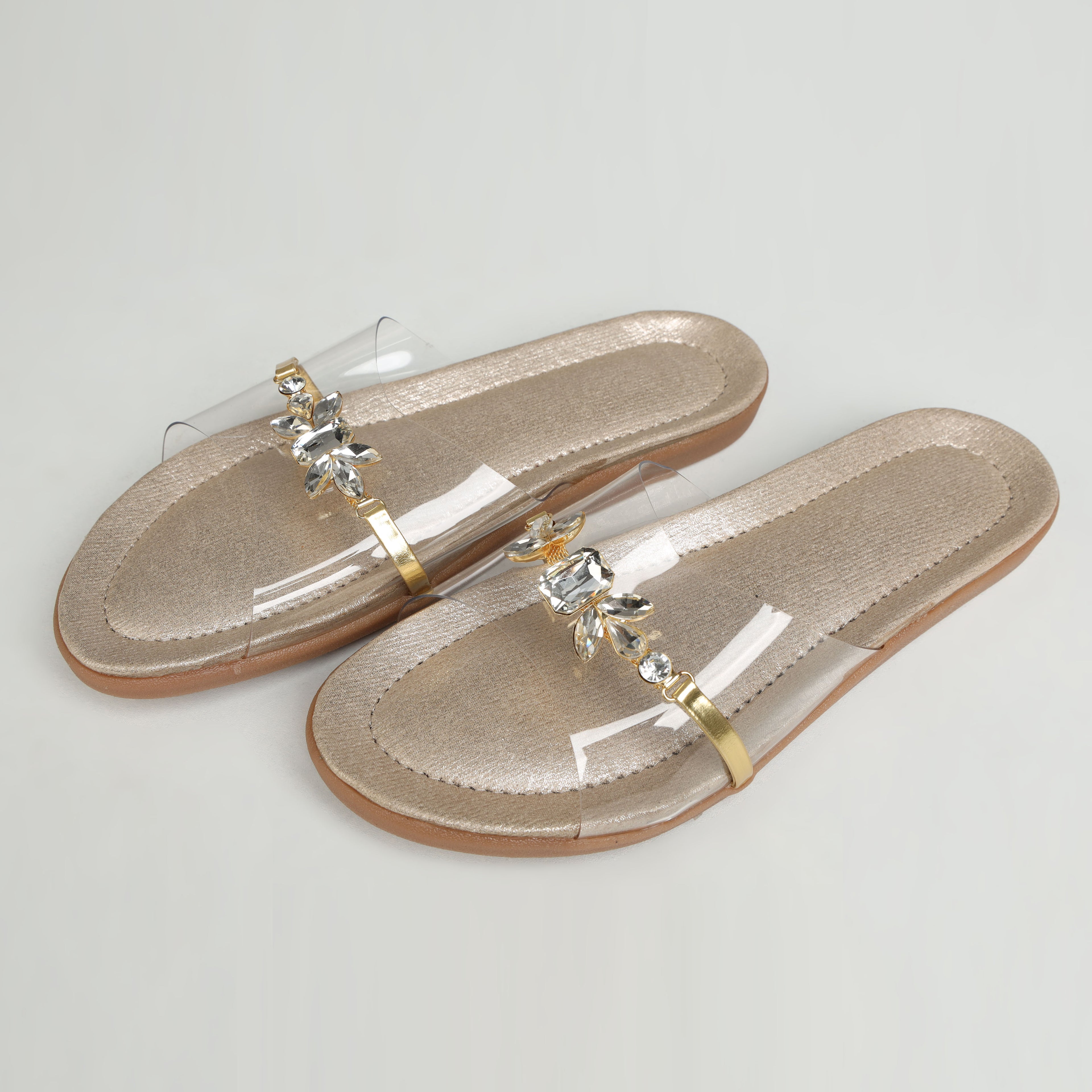 Pair of Riwaaz Ira dull gold crystal transparent strap flats – angled product shot on grey background showing rubber sole and gold metal toe-post detail