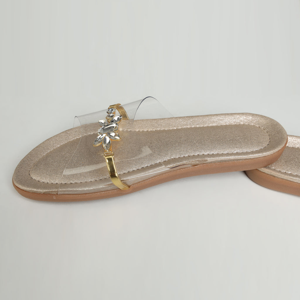 Pair of Riwaaz Ira dull gold crystal-embellished transparent silicone strap flats – product shot on cream surface with white vase and gold geometric tray in background