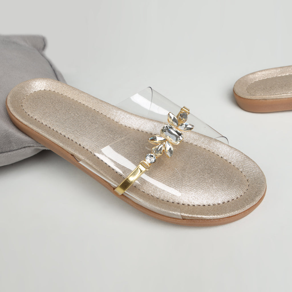 Model wearing Riwaaz Ira dull gold crystal transparent strap flats with cream embroidered salwar – close-up foot shot on stone floor in Indian courtyard setting