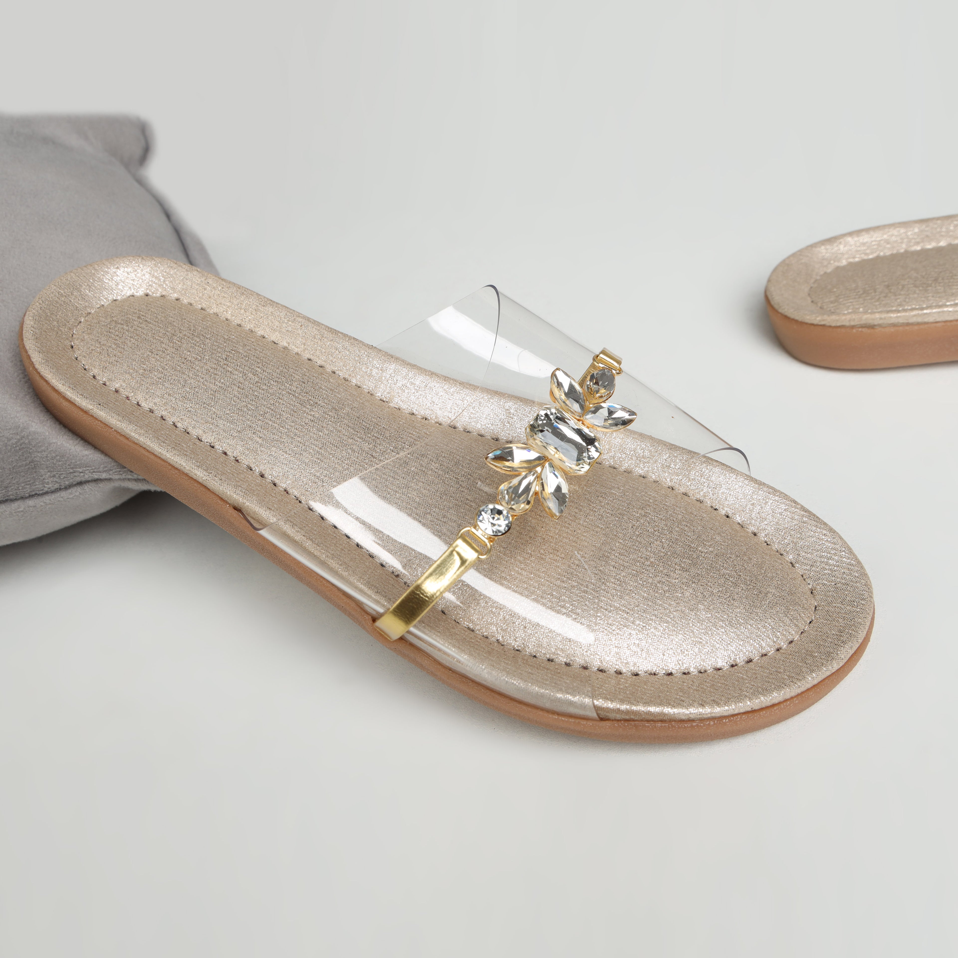Model wearing Riwaaz Ira dull gold crystal transparent strap flats with cream embroidered salwar – close-up foot shot on stone floor in Indian courtyard setting