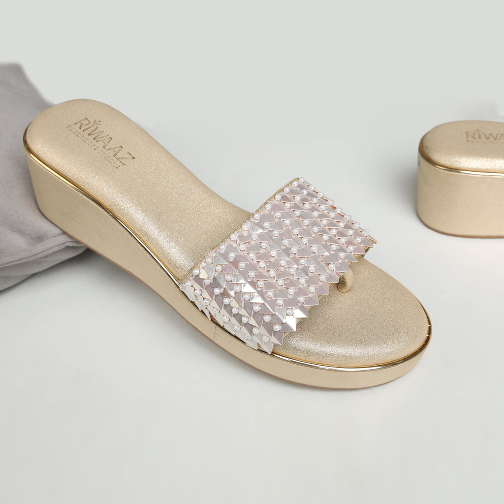 Model wearing Riwaaz Chandni Glow dull gold pearl and sequin wedge sandals with cream embroidered salwar – close-up foot shot on jute rug in warm Indian interior setting