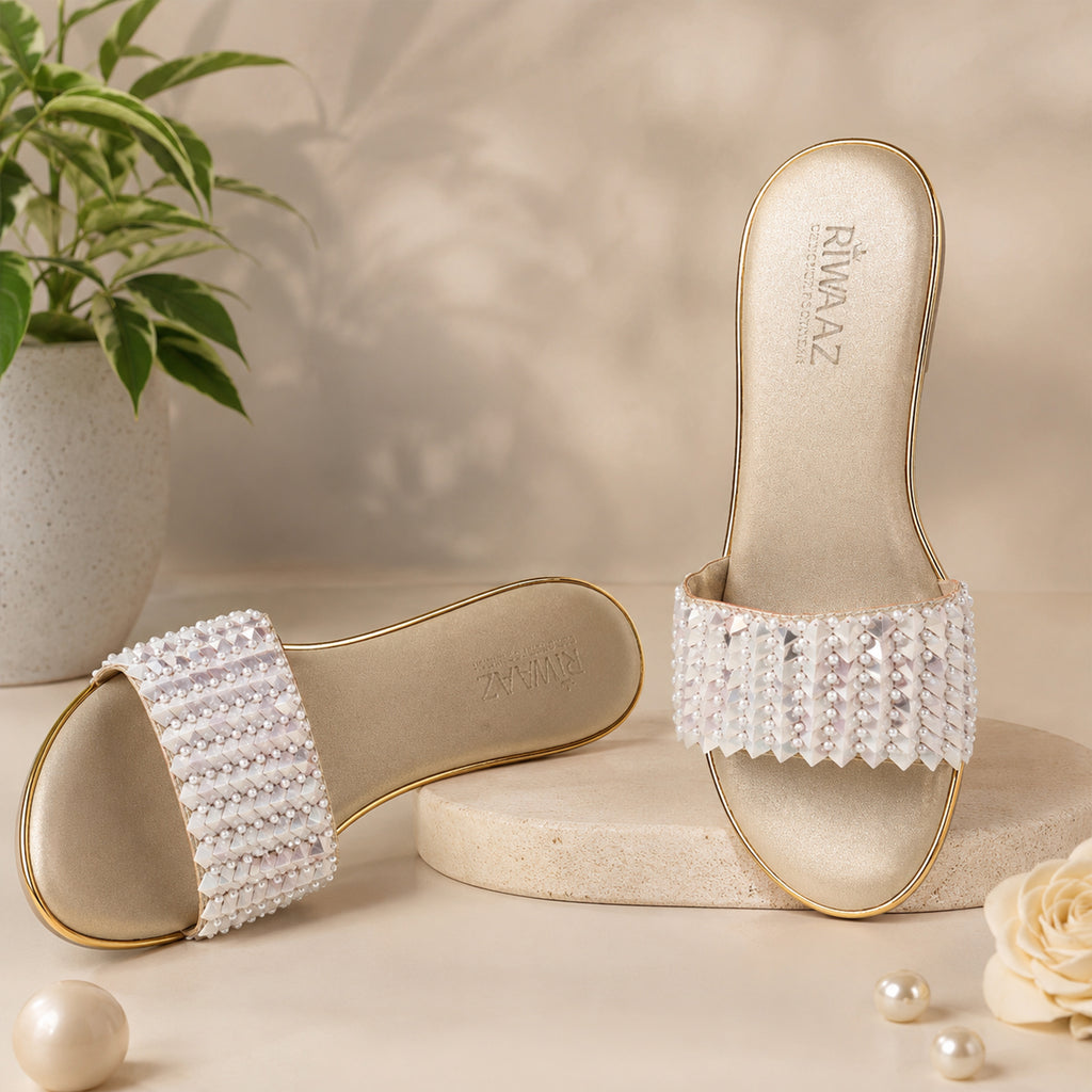 Model seated in cream embroidered kurta set wearing Riwaaz Chandni Glow dull gold pearl and sequin wedge sandals – full lifestyle shot in warm Indian interior with rattan chair and pink flowers