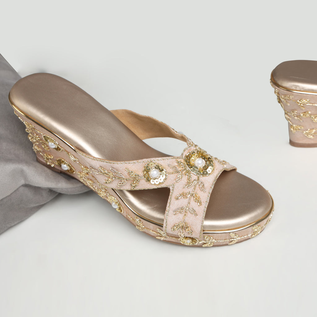 Model wearing Riwaaz Noor rose gold zari embroidered bridal wedge heels with peach embroidered salwar – close-up foot shot showing pearl sequin cross-strap and embroidered wedge on marble floor