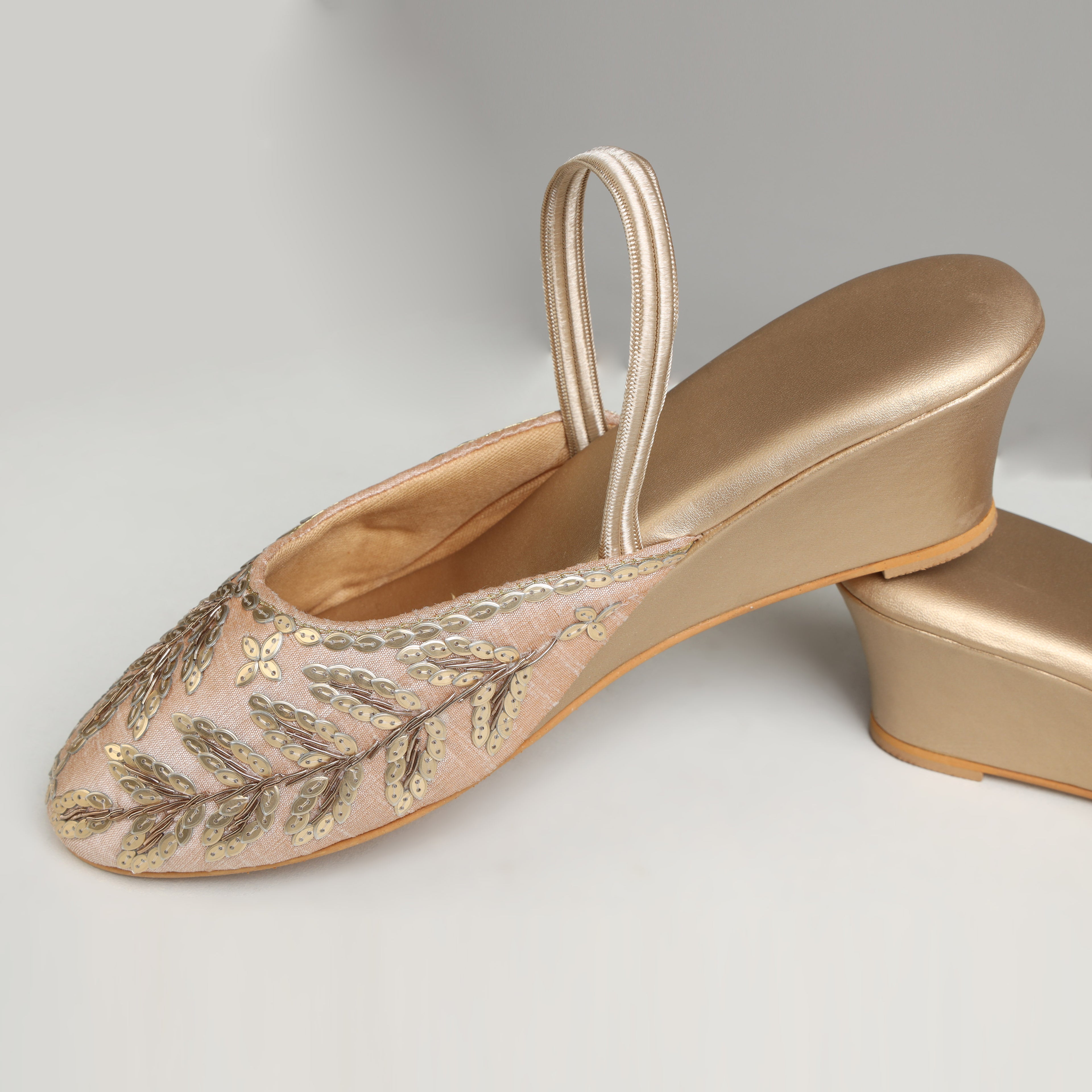 Pair of Riwaaz Zehra dull gold sequin leaf embroidered slingback wedge mules – lifestyle flat lay on cream satin surface with gypsophila flowers, scattered pearls and brass bowl in warm Indian setting