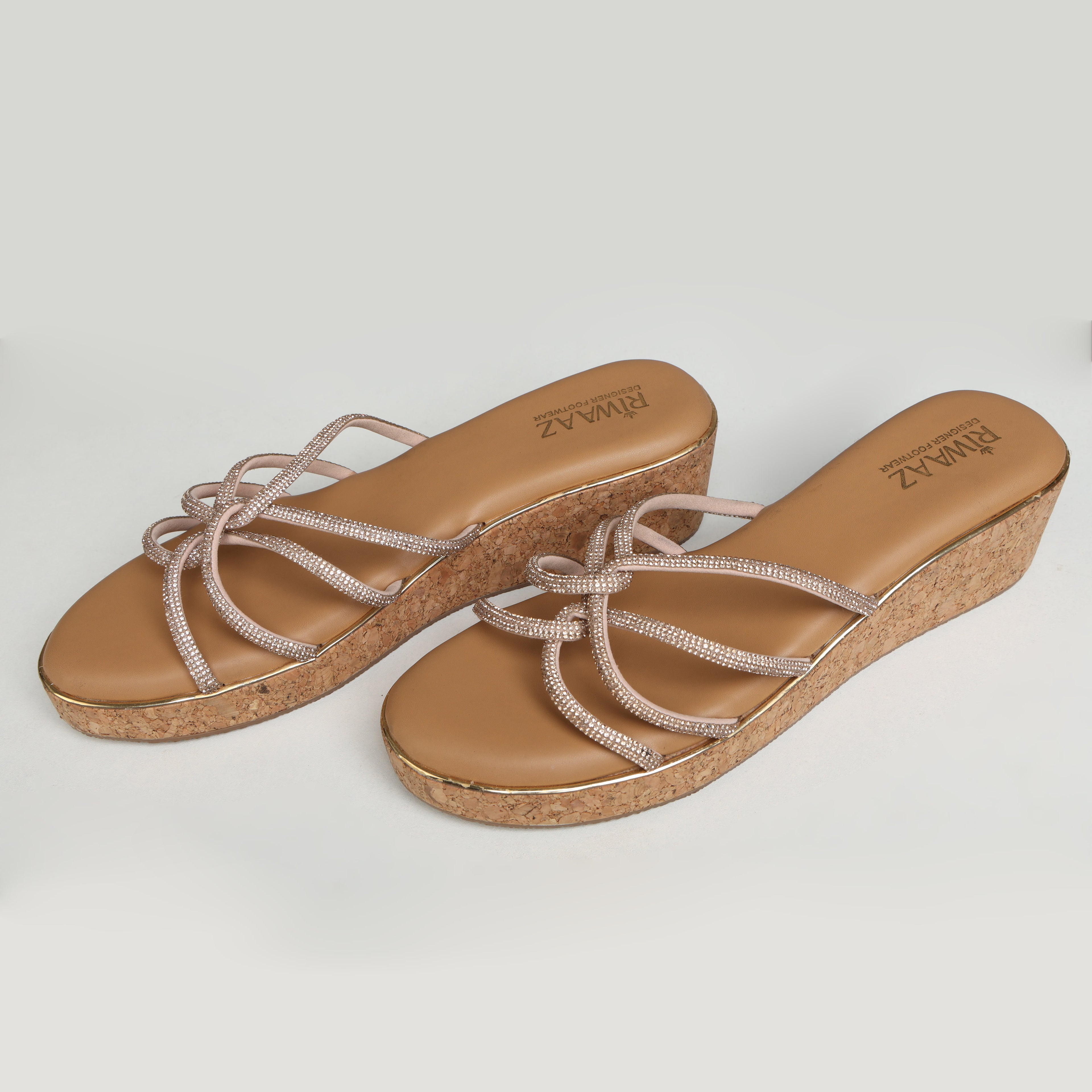 Pair of Riwaaz Zarina Glow dark beige crystal crossover cork wedge platforms on grey background – angled three-quarter view showing full rhinestone crossover strap detail and natural cork wedge with gold trim outsole
