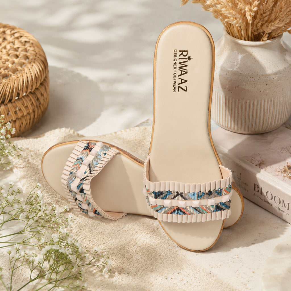 Model seated in ivory floral embroidered kurta set wearing Riwaaz Elara cream printed pleated strap wedge slides – full lifestyle shot in warm Indian interior with large ceramic vase and gypsophila flowers